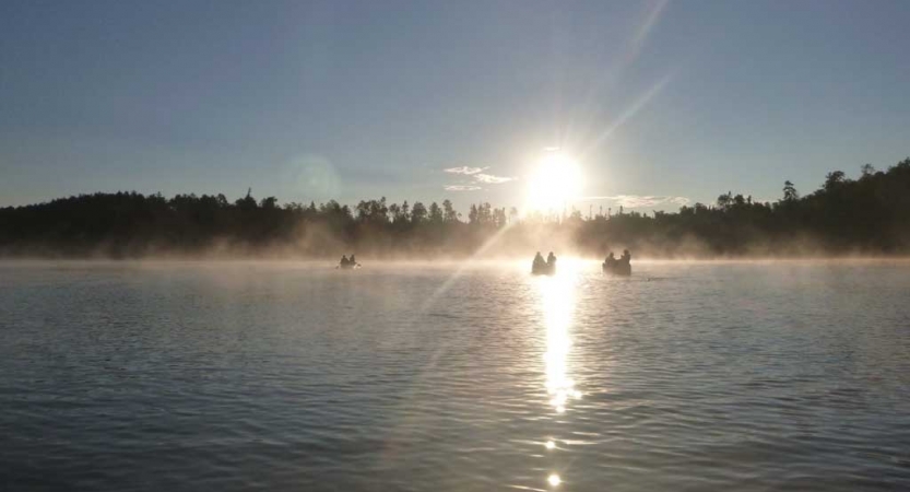 In the distance, three canoes are paddled toward the sun, through fog sitting on the water. 